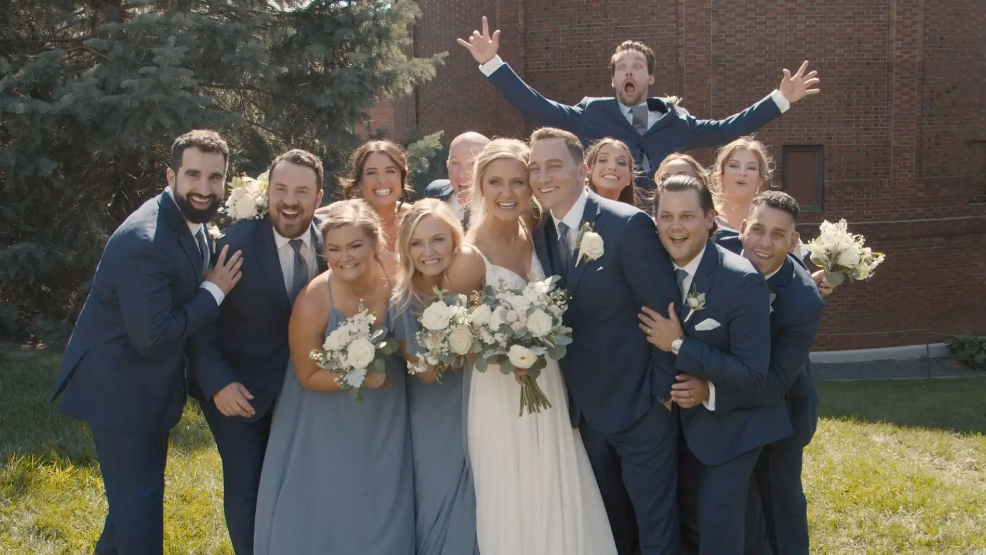 Candid moment captured by the wedding videographer of Bethany, Nick, and their wedding party in Grand Rapids, Michigan. The group is joyfully posing with smiles and bouquets, set against lush Michigan greenery.