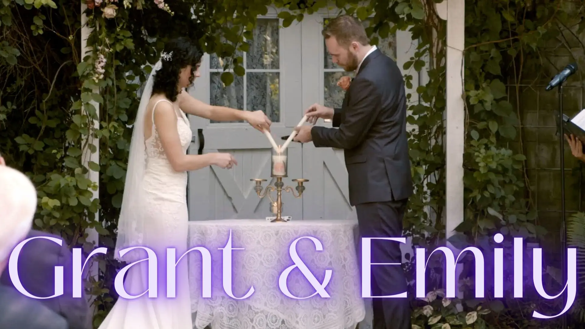 Bride Emily and groom Grant light a unity candle during their wedding ceremony at The Blue Dress Barn in Benton Harbor, Michigan. The photo has a romantic, candid feel, with "Grant & Emily" written in light purple text at the bottom.
