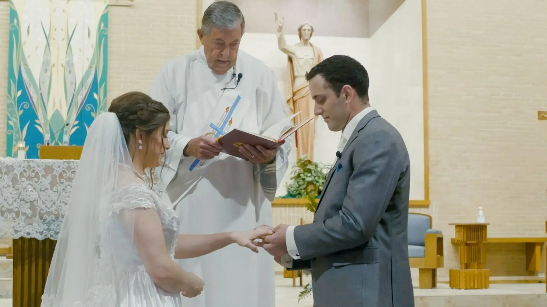 Elizabeth and Aaron exchange rings during their wedding ceremony at Sacred Heart Catholic Church in Mt Pleasant on July 15, 2023. The priest stands behind them, holding a book and officiating.