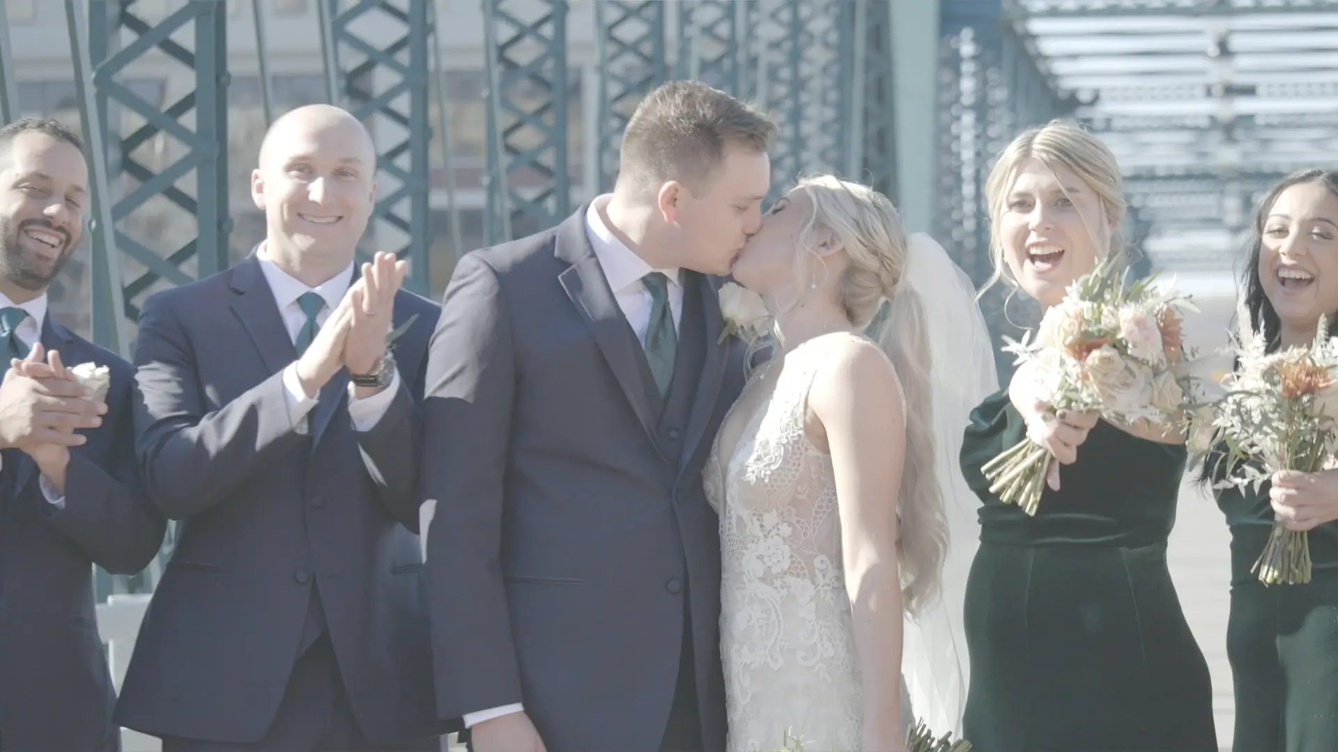 Dana and Mason share a kiss on a bridge in Grand Rapids, Michigan. Surrounded by their joyous wedding party, the bridesmaids and groomsmen smile and clap, celebrating the newlyweds in their fall-themed wedding.