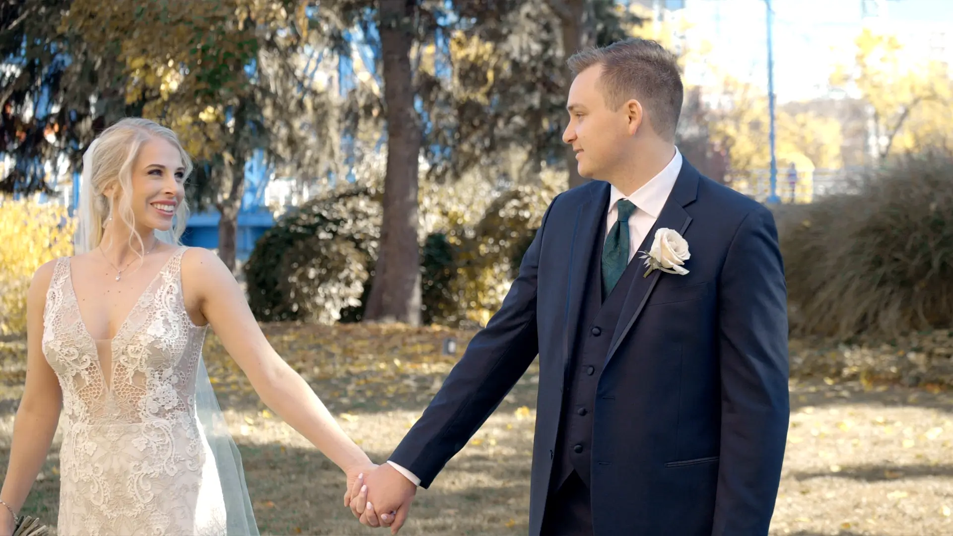 Dana and Mason are smiling at each other and holding hands outdoors on their wedding day in Grand Rapids, Michigan. Dana wears a white lace dress, and Mason is in a dark suit with a white rose. Colorful autumn foliage is visible in the background.