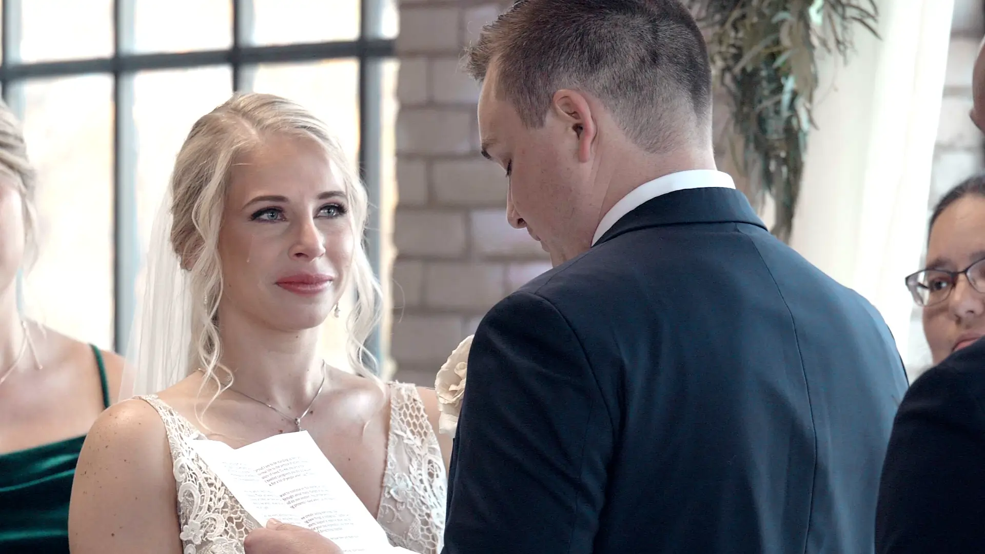 Bride Dana looks emotional with a tear on her cheek as she shares a sentimental moment with groom Mason during their Jewish wedding ceremony at Studio D2D. The groom is reading from a piece of paper, and part of the wedding party is visible.