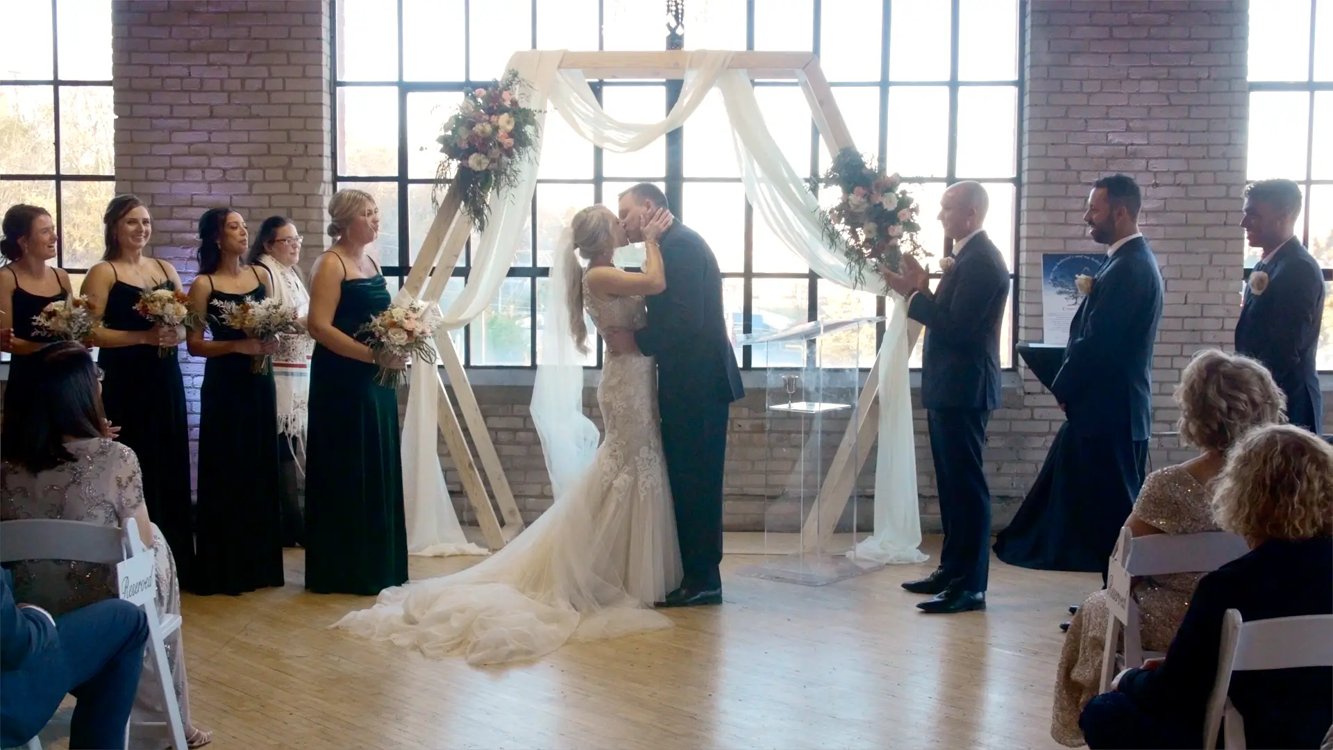 Bride and groom, Dana and Mason, share a kiss under an arch adorned with flowers and draped fabric during their wedding ceremony. Bridesmaids and groomsmen in formal attire stand on either side, while guests watch from seated rows.