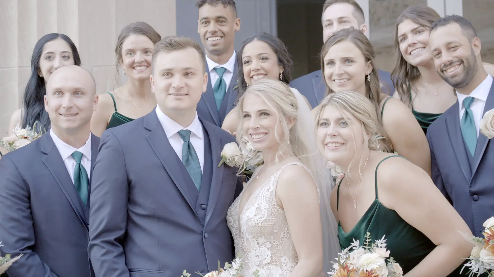 Bride Dana and groom Mason are joyfully for their wedding videographer outside a Grand Rapids venue. Everyone is dressed elegantly, with the bride in a detailed lace gown and the groom in a dark suit with a white rose boutonnière.