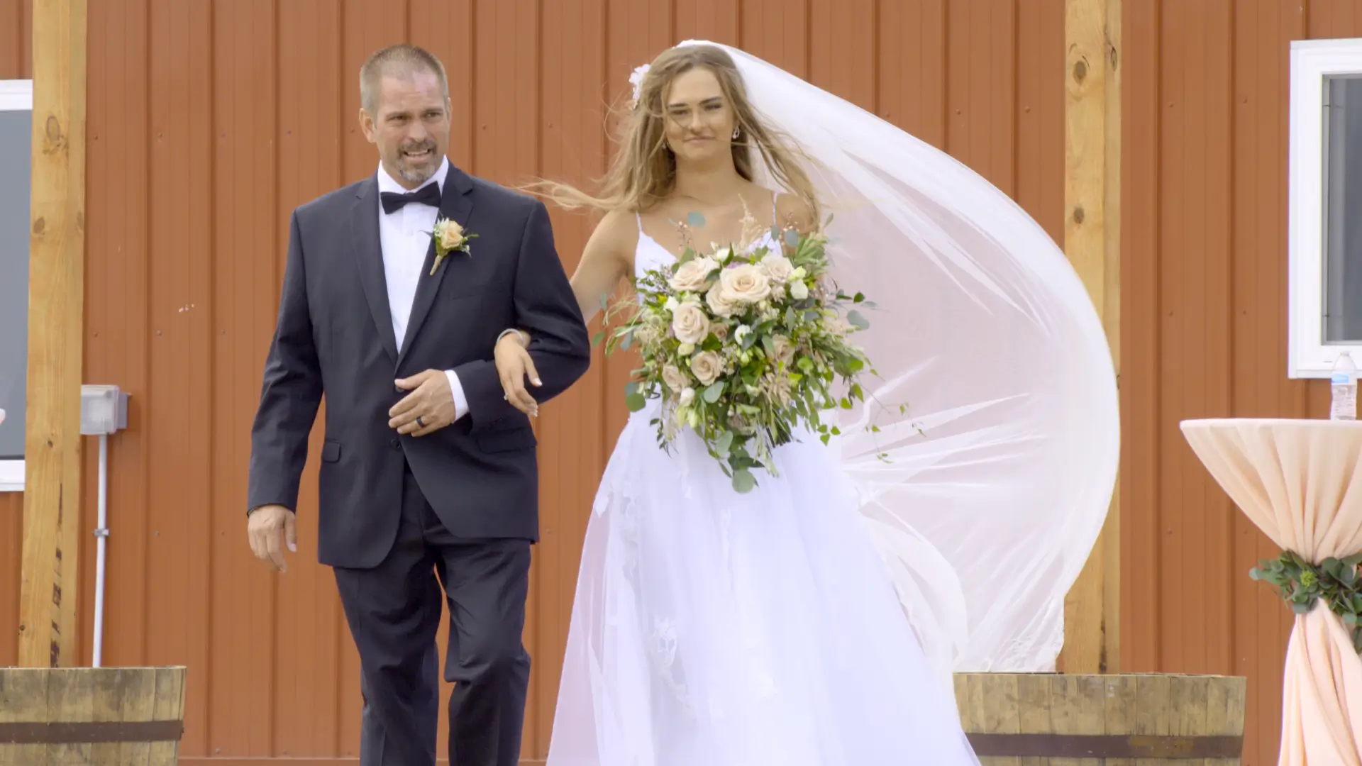 The bride, Mikayla, walks down the aisle with her father at Apple Valley Event Center in Sparta, Michigan. She wears a white gown and holds a bouquet of flowers, while her long veil flows behind. They stand against a rustic red backdrop.