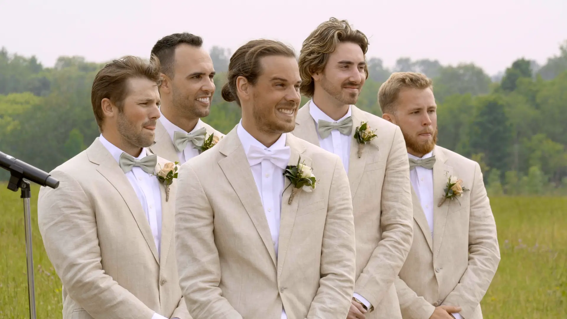 Groom and four groomsmen in light beige suits with green bow ties, standing outside at Apple Valley Event Center. They are smiling with lush greenery in the background.
