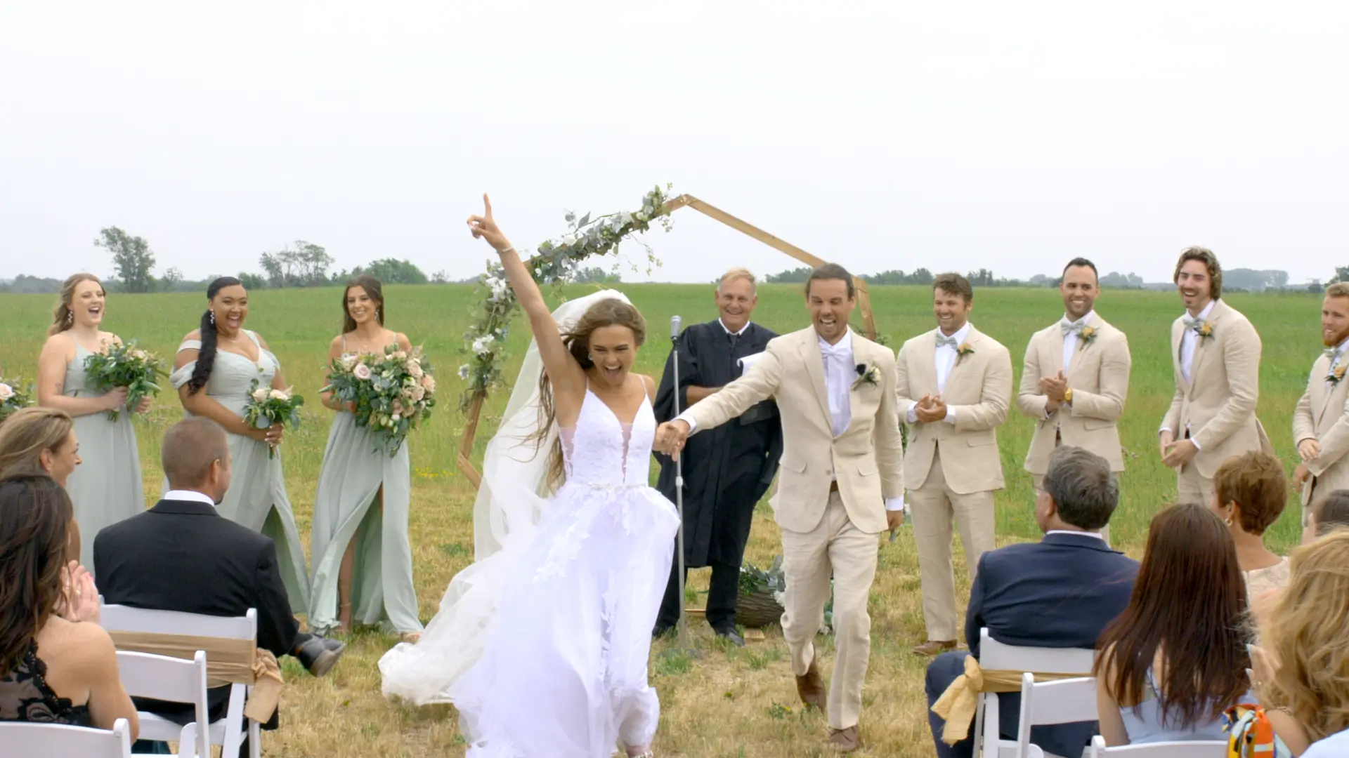 Bride Mikayla and groom Jackson celebrate joyously after their wedding ceremony at Apple Valley Event Center in Sparta, Michigan. Their wedding party stands beside them, smiling and clapping in the lush green fields.