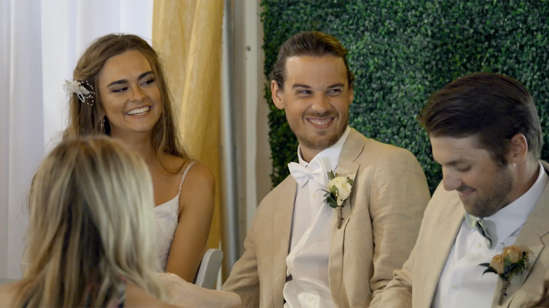 Bride Mikayla and groom Jackson are smiling and seated at their wedding reception at the Apple Valley Event Center. They are dressed in light-colored attire, with greenery serving as a backdrop. Another man in similar attire is sitting beside them.