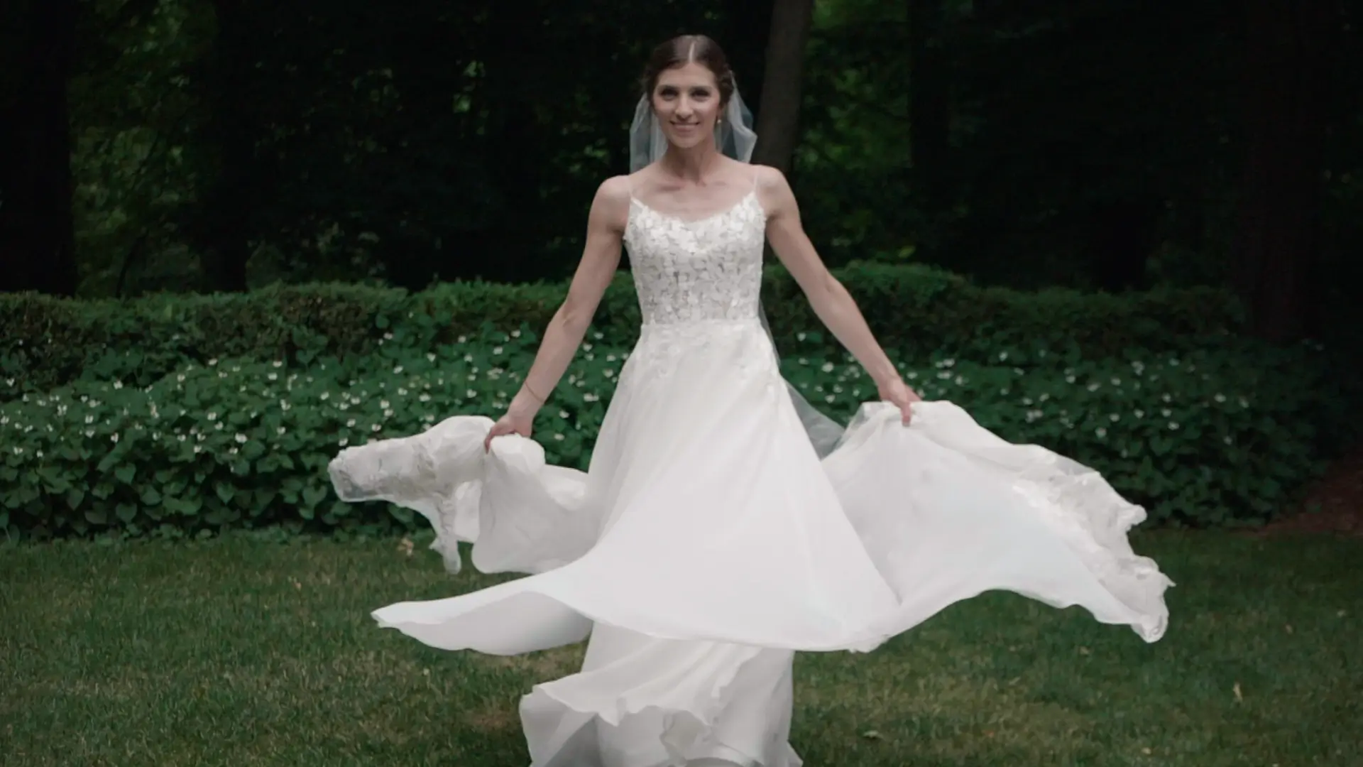 A bride, Kathryn, twirling in her white wedding dress, smiling and holding the sides of her gown, against a backdrop of lush, Michigan greenery.