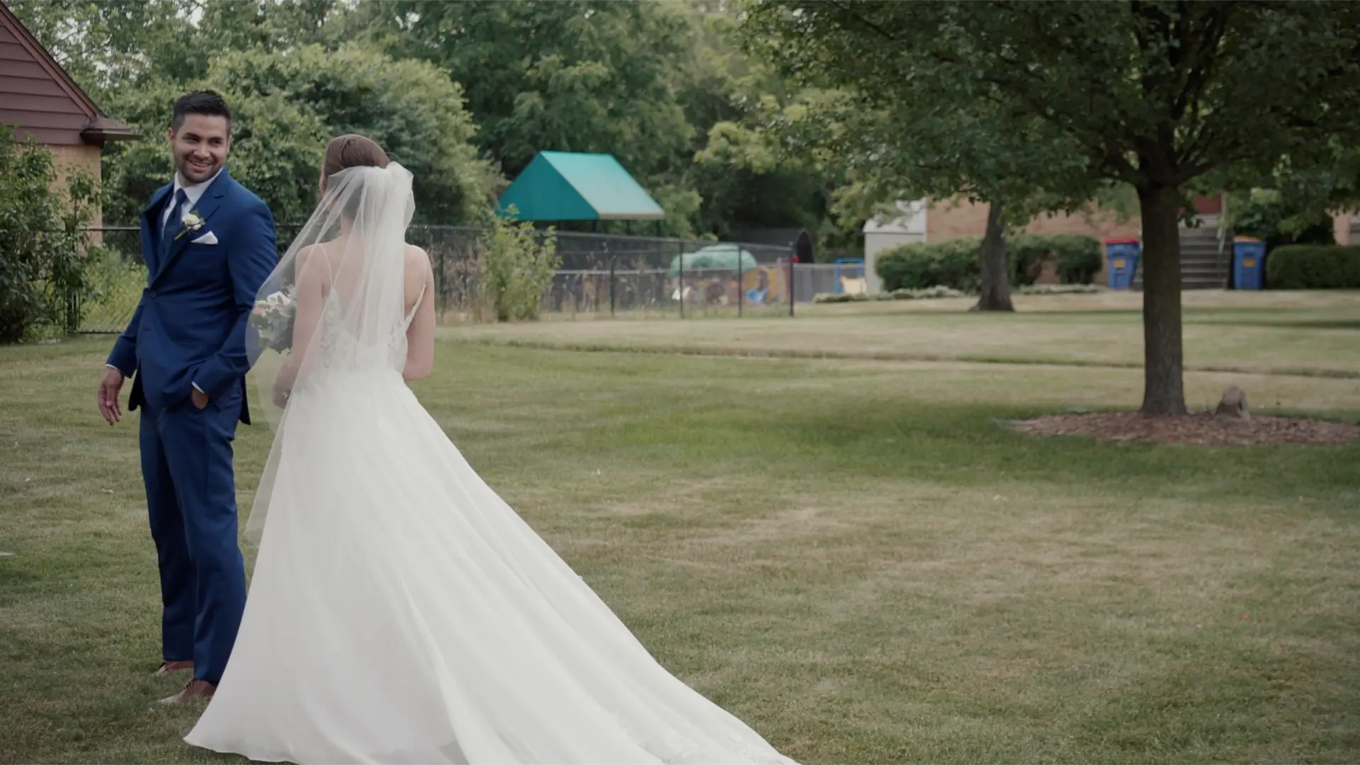 The groom, Esteban, in a blue suit, smiles at his bride, Kathryn, in a white wedding dress and veil, as they stand in a grassy area outside their church in downtown Grand Rapids. Trees and a playground are visible in the background.