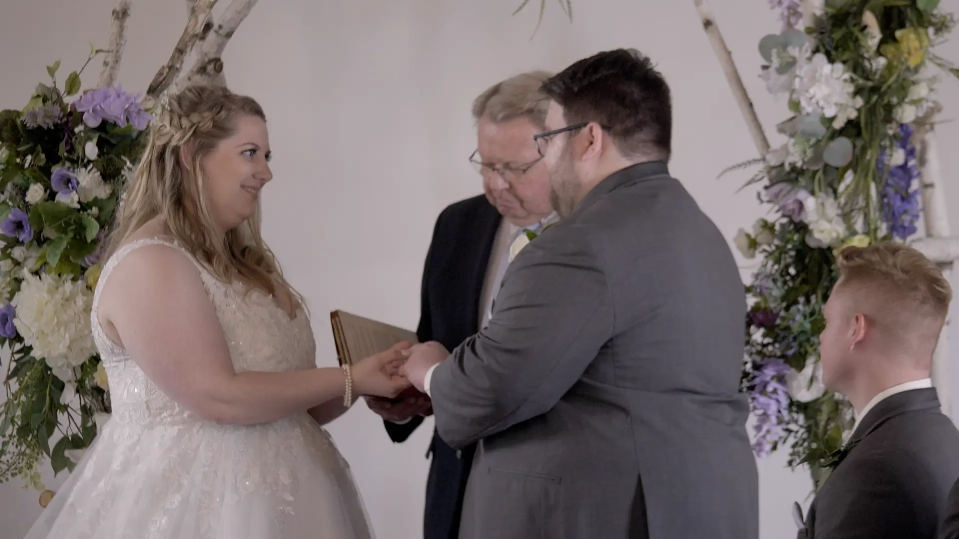 Bride Amanda and groom Clark exchange vows in front of an officiant, surrounded by floral arrangements in a church setting. Amanda wears a white lace dress, while Clark is in a gray suit. Another man, part of their wedding party, sits to the right.