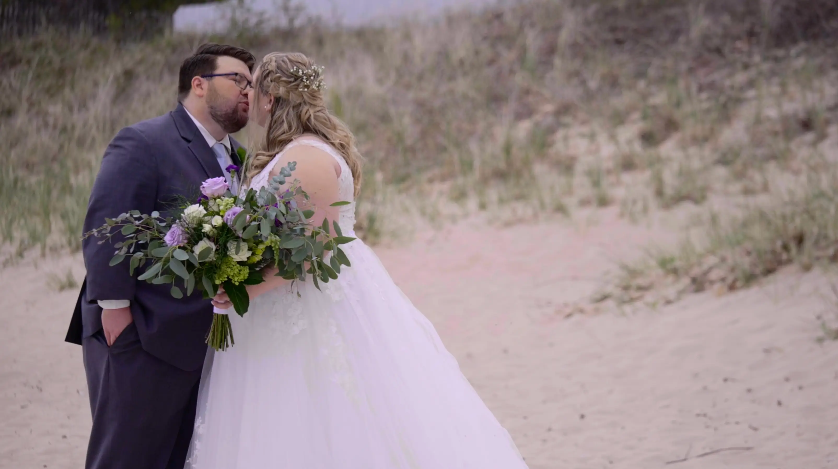 In this snapshot from their wedding highlight, bride Amanda and groom Clark share a kiss on a sandy beach in Traverse City, Michigan, surrounded by Michigan grass dunes. Amanda holds a lush bouquet, and both are dressed in wedding attire.
