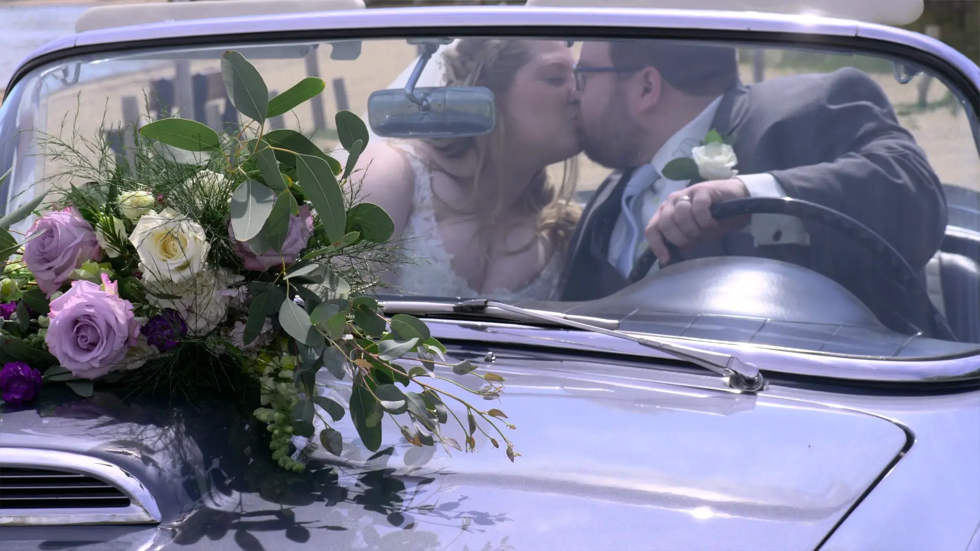 Amanda and Clark kiss in a vintage car on their wedding day in Traverse City. The car's windshield showcases a bouquet of purple and white roses.
