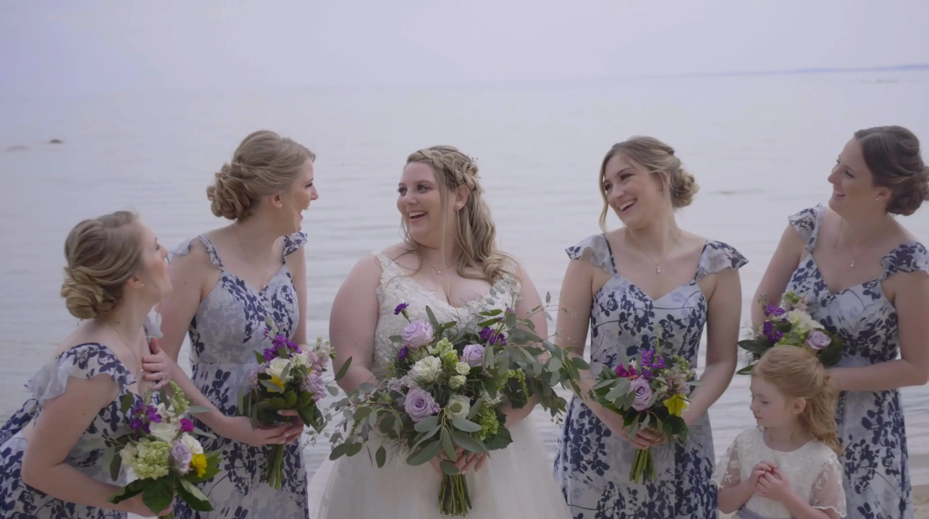 In this still from their wedding highlight video, bride Amanda shares a joyful moment with her bridesmaids by the serene lakeside in Traverse City, Michigan. The bridesmaids wear floral-patterned dresses and hold bouquets as the flower girl looks on.