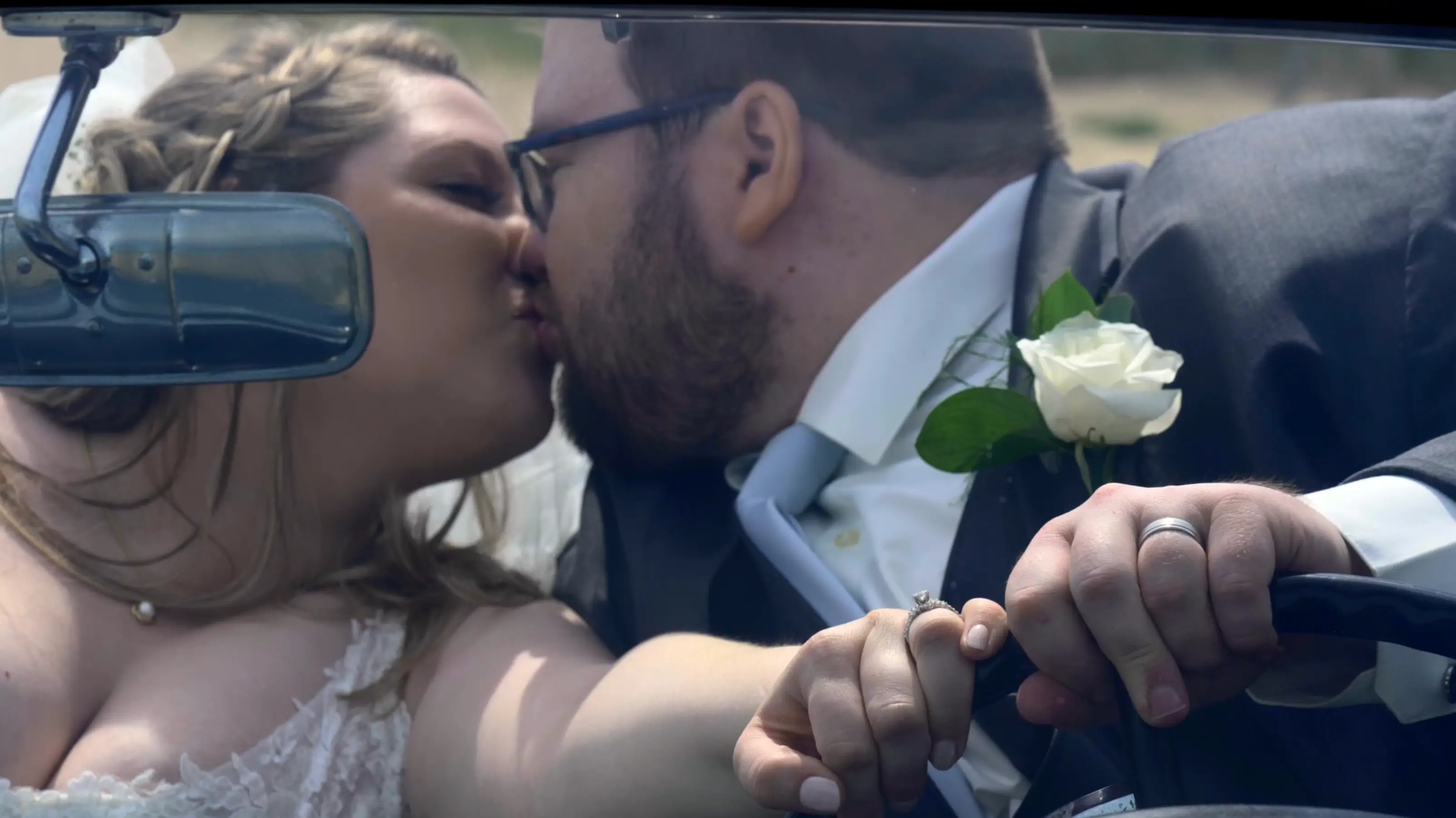 Amanda and Clark share a kiss inside a car, holding hands on the steering wheel. Amanda wears a lace wedding dress and shows her engagement ring, while Clark is in a suit with a white boutonniere. The rearview mirror partially obscures their view.