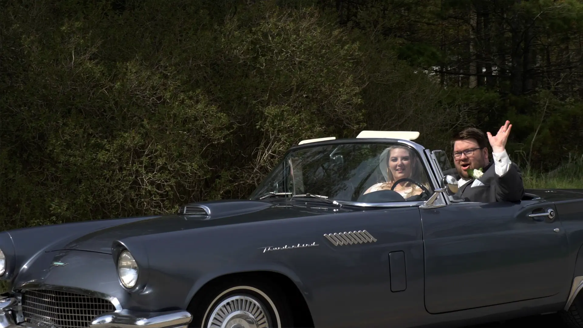 A bride and groom are sitting in a classic convertible car. The bride, Amanda, is driving, and the groom, Clark, is waving cheerfully. The couple seems happy as they celebrate their wedding in Traverse City, Michigan.