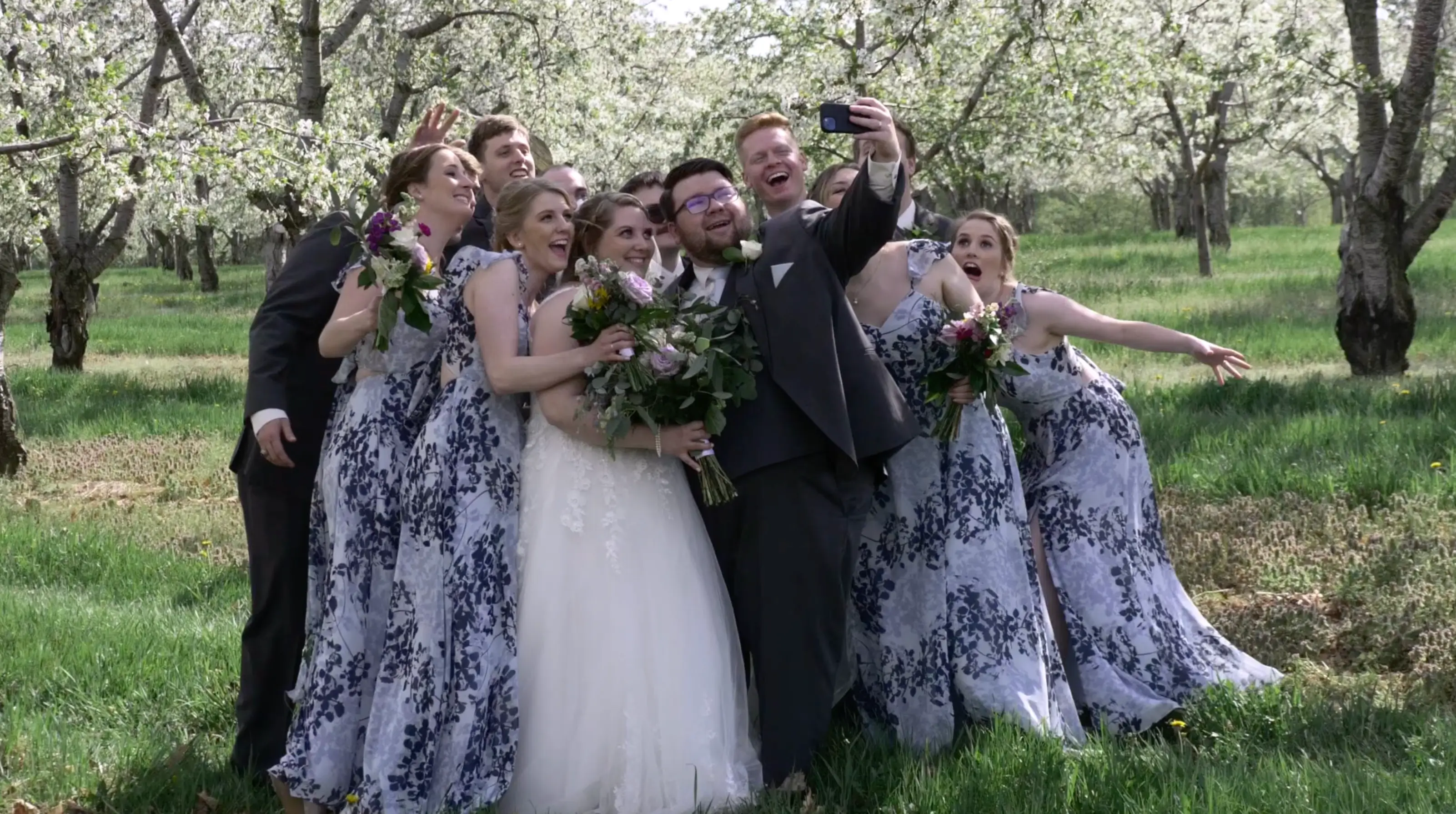Bride Amanda and groom Clark take a group selfie with their bridesmaids and groomsmen surrounded by blooming Michigan cherry trees in a Traverse City orchard. The bridesmaids wear blue floral dresses, and everyone is smiling and holding bouquets.