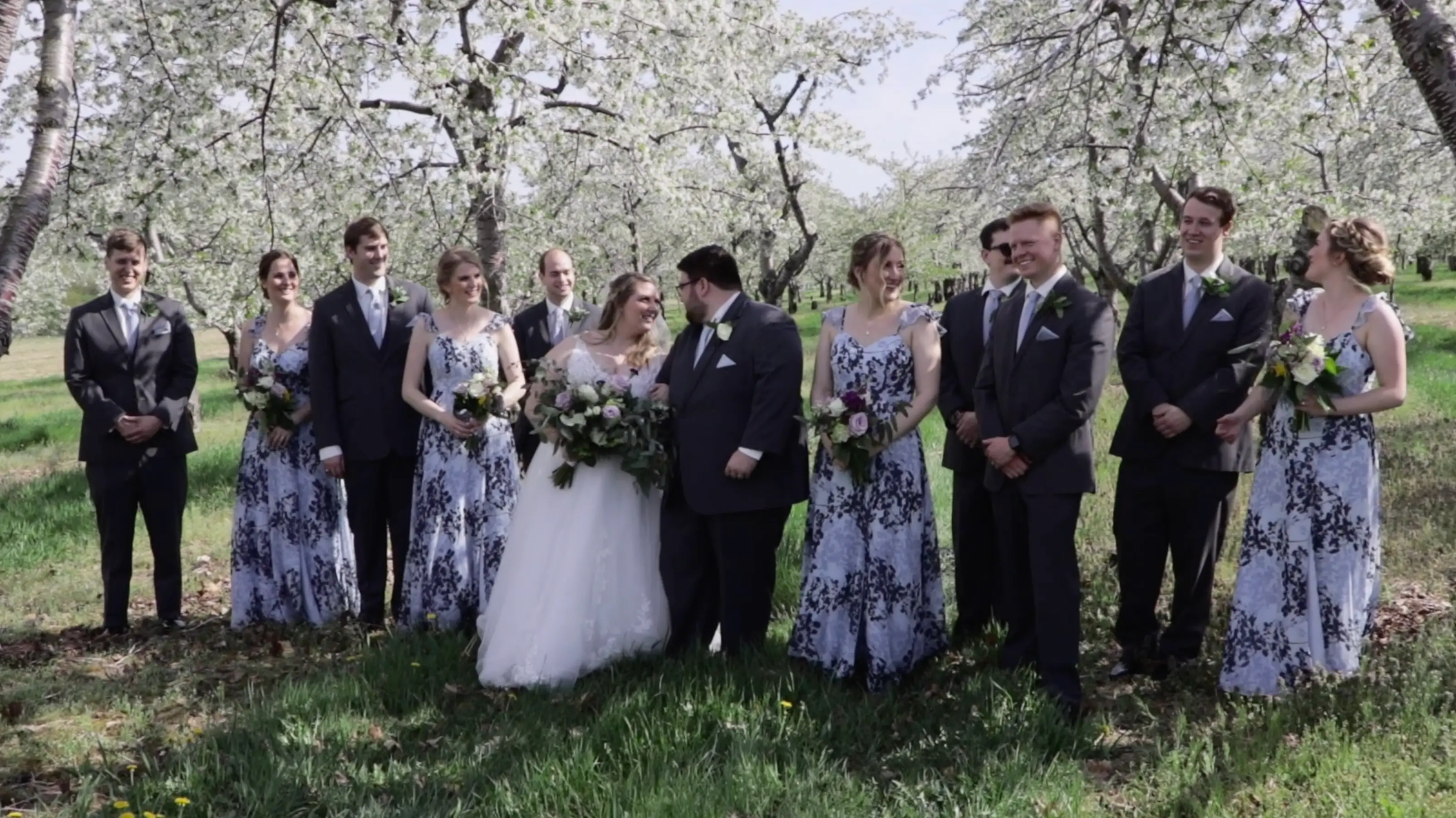 In this still from their wedding video in Traverse City, Michigan, Amanda and Clark share a moment with their bridal party in a blooming cherry orchard. The bridal party is dressed in dark suits and floral dresses, holding bouquets of flowers.