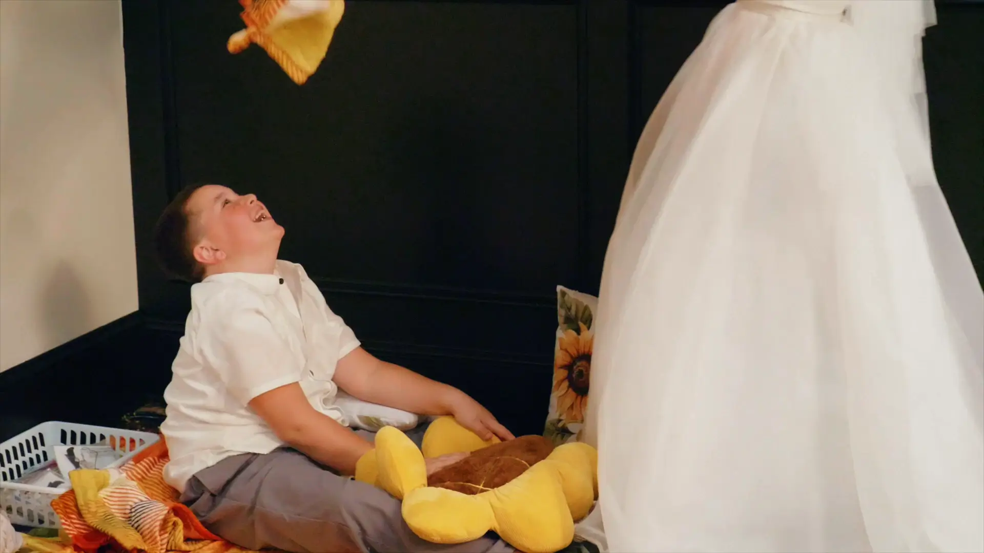 In this candid moment captured by the wedding videographer, a young boy joyfully laughs while sitting on the floor, surrounded by playful Michigan-themed decor, as a woman in a wedding dress plays with him.