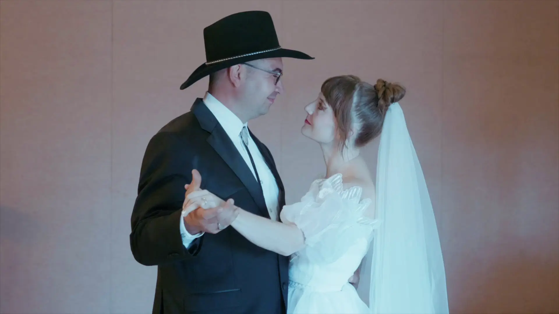 Bride Felicia and groom Leigh share a romantic first dance at their wedding reception. Felicia wears a white gown and veil, while Leigh dons a black suit with a cowboy hat. The image captures their loving gaze in Howell at Crystal Gardens Event Center.