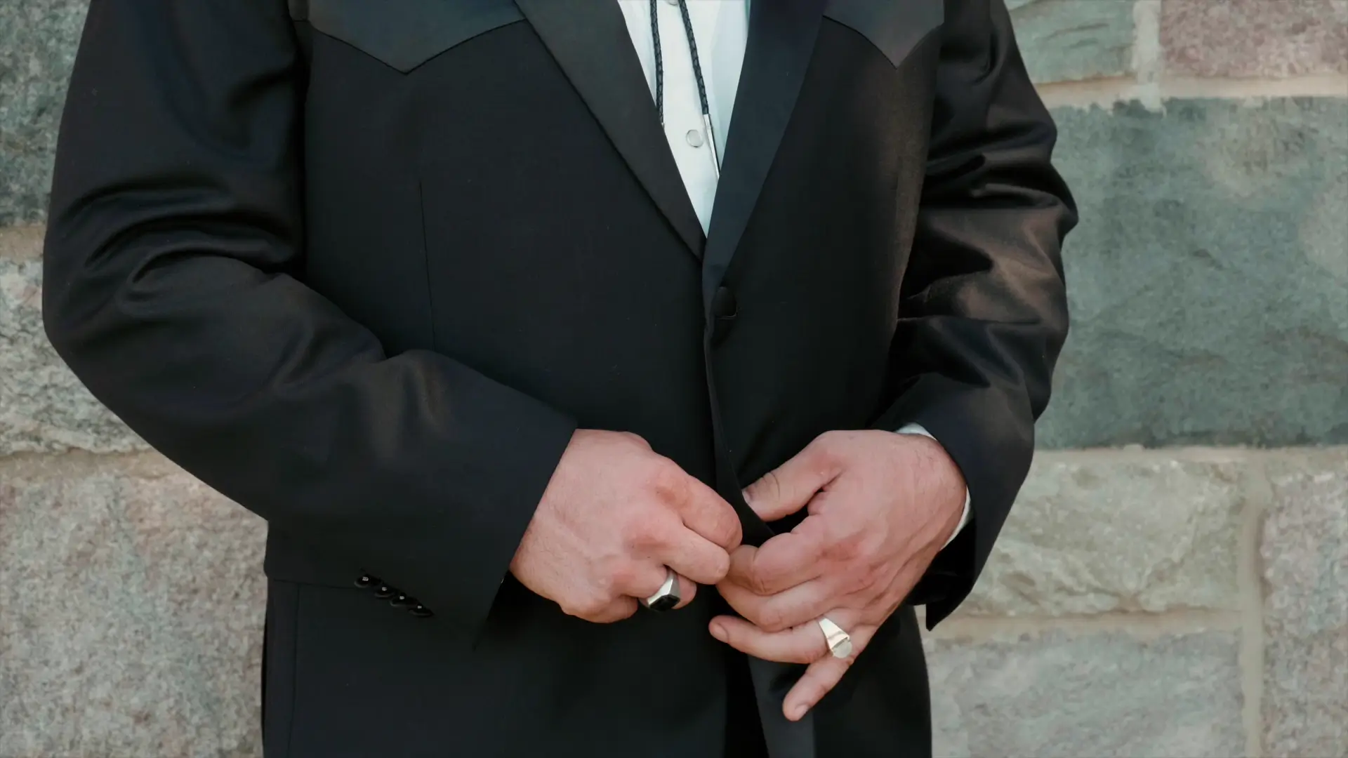 A close-up of the groom in his black suit, adjusting his jacket in front of a stone wall, showcasing his elegant attire before his Michigan wedding.