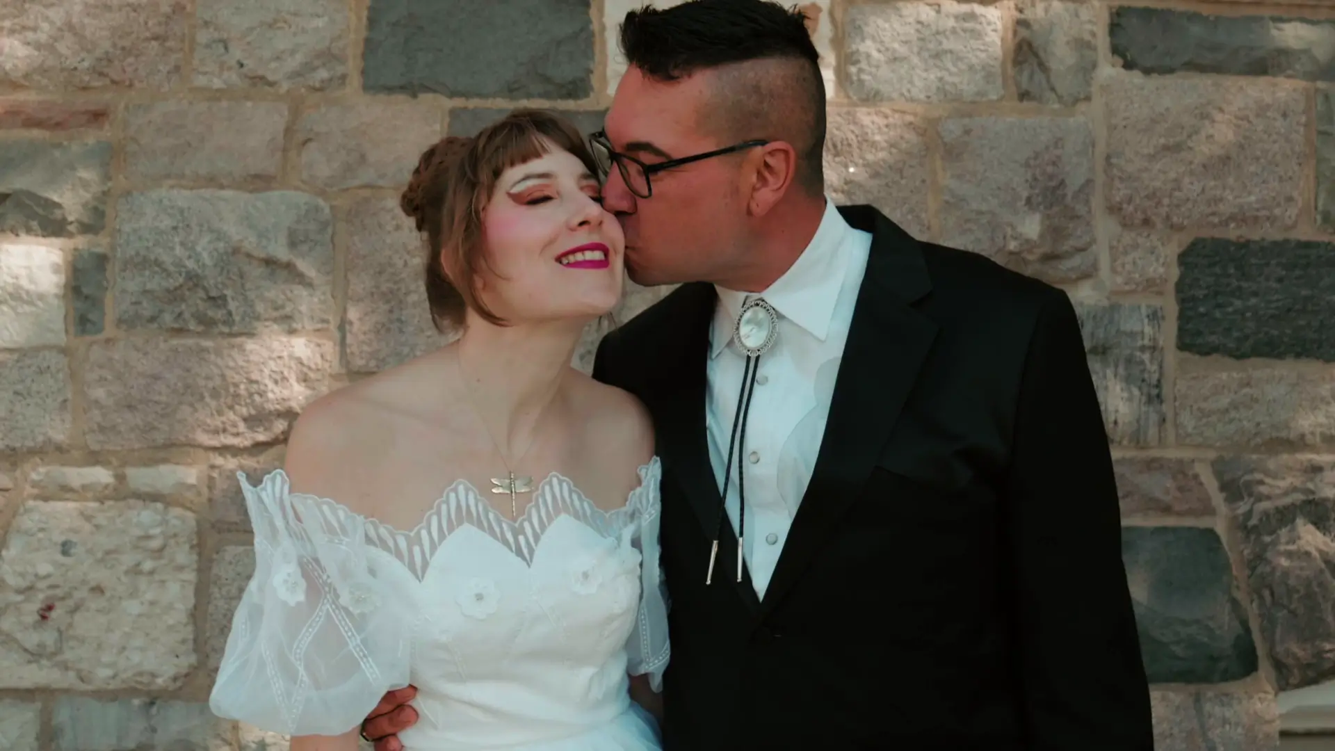 Bride Felicia and groom Leigh share a joyful moment, with Leigh kissing Felicia on the cheek. Felicia is wearing an off-the-shoulder white wedding dress, and Leigh is dressed in a black suit with a bolo tie. They stand against a stone wall backdrop.