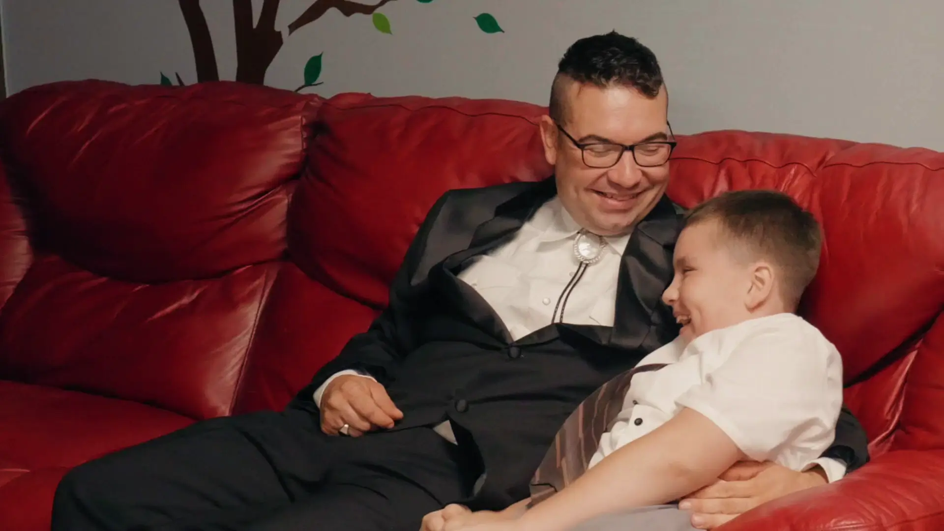 In this candid moment captured by the wedding videographer, the groom and a child share a joyful laugh on a red couch, embodying the heartwarming and family-focused atmosphere of Felicia and Leigh's wedding in Michigan.
