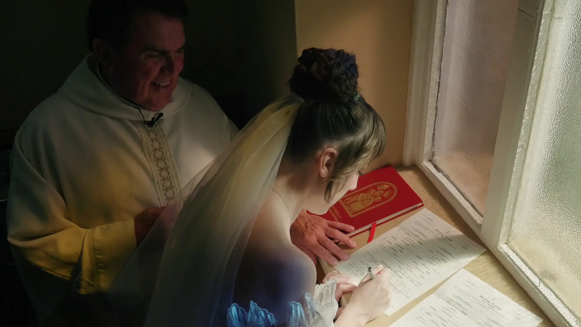 Bride Felicia, in a white dress and veil, signs marriage documents at a window-lit table in St. Augustine Catholic Church, Howell, Michigan. A smiling priest stands nearby observing the moment.