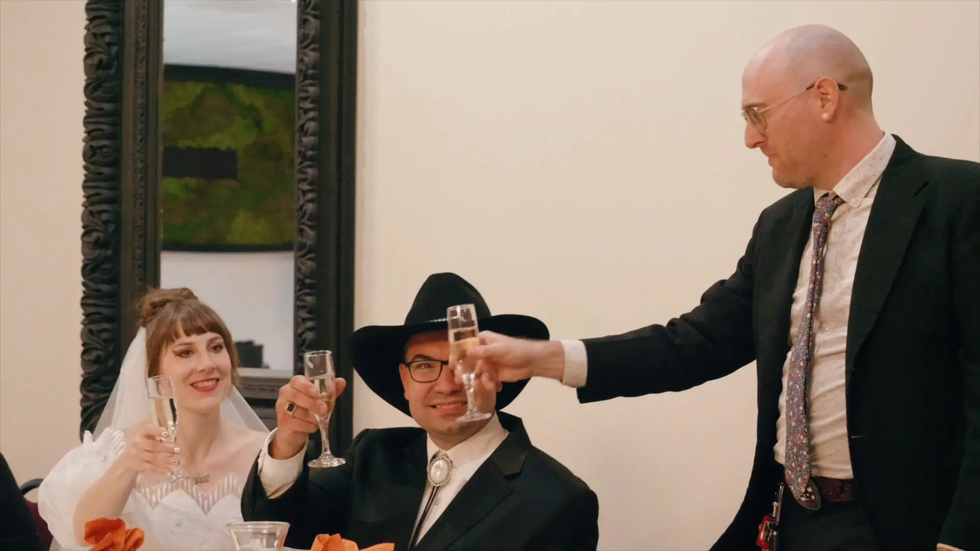 Bride Felicia and groom Leigh toast with a guest at their wedding reception in Howell, Michigan. Felicia is smiling in a wedding dress, Leigh wears a black cowboy hat and glasses, and the guest in a suit raises a glass, all appearing joyful.