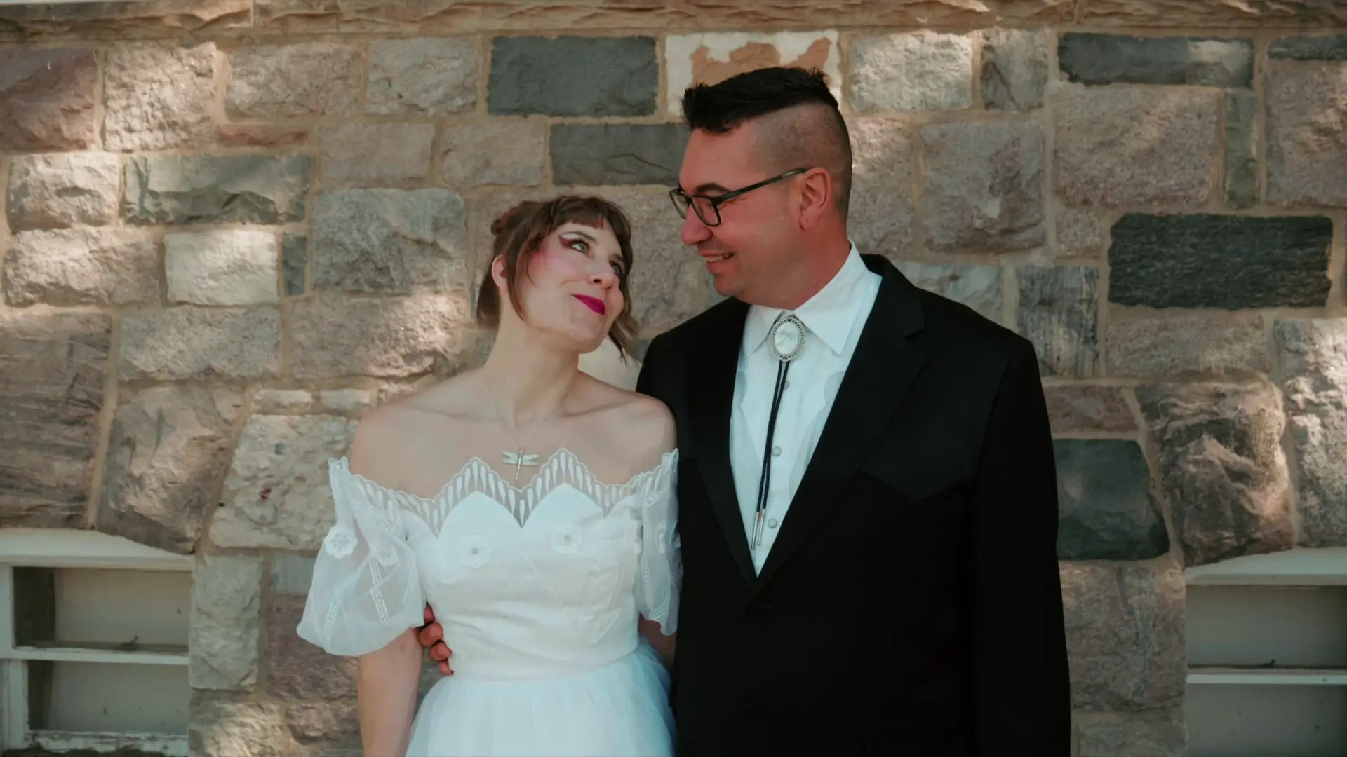 Bride and groom smiling at each other in front of a stone wall. The bride wears an off-the-shoulder lace dress and the groom is in a black suit with a bolo tie. The setting suggests a warm, intimate moment on their wedding day.