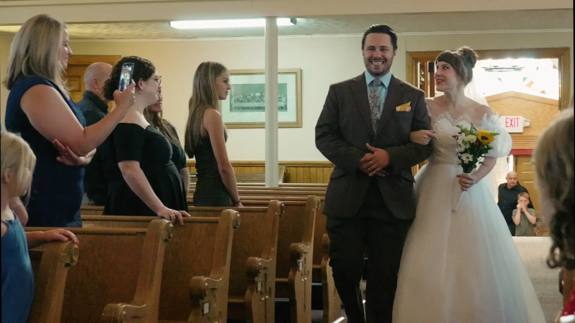 A bride is escorted down the aisle by a man in a gray suit, who playfully makes a silly face during Felicia & Leigh's Catholic wedding ceremony. The bride holds a bouquet of sunflowers and daisies.