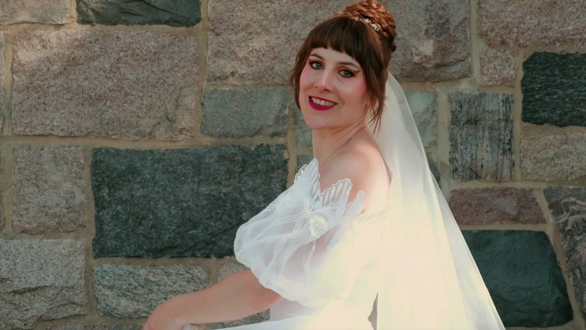Bride Felicia smiles in her wedding gown with a lace veil, twirling in front of a stone wall.
