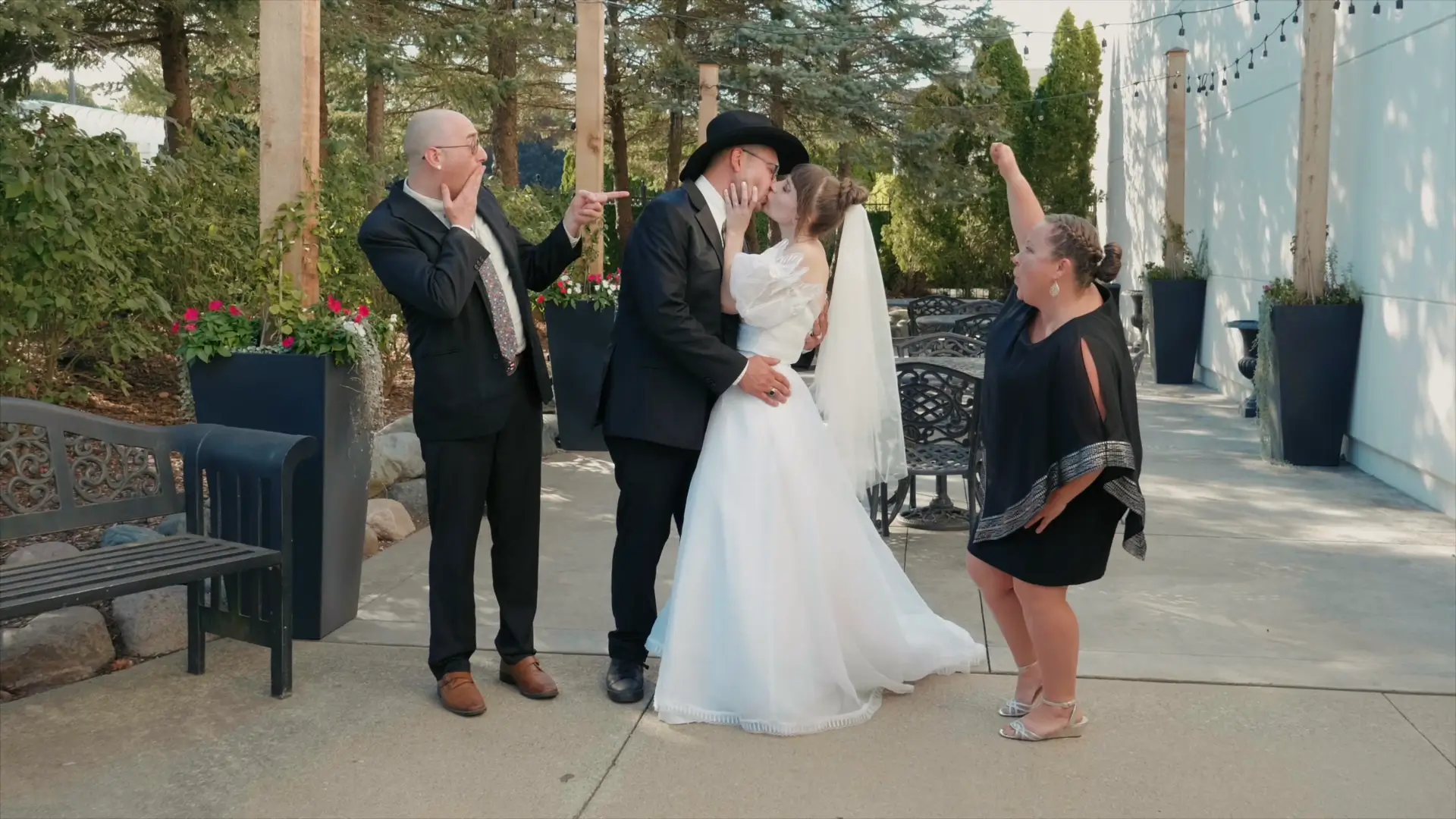 Bride and groom share a kiss outdoors in Howell, Michigan, while two excited onlookers react happily. The lush greenery and festive atmosphere set a joyful scene.