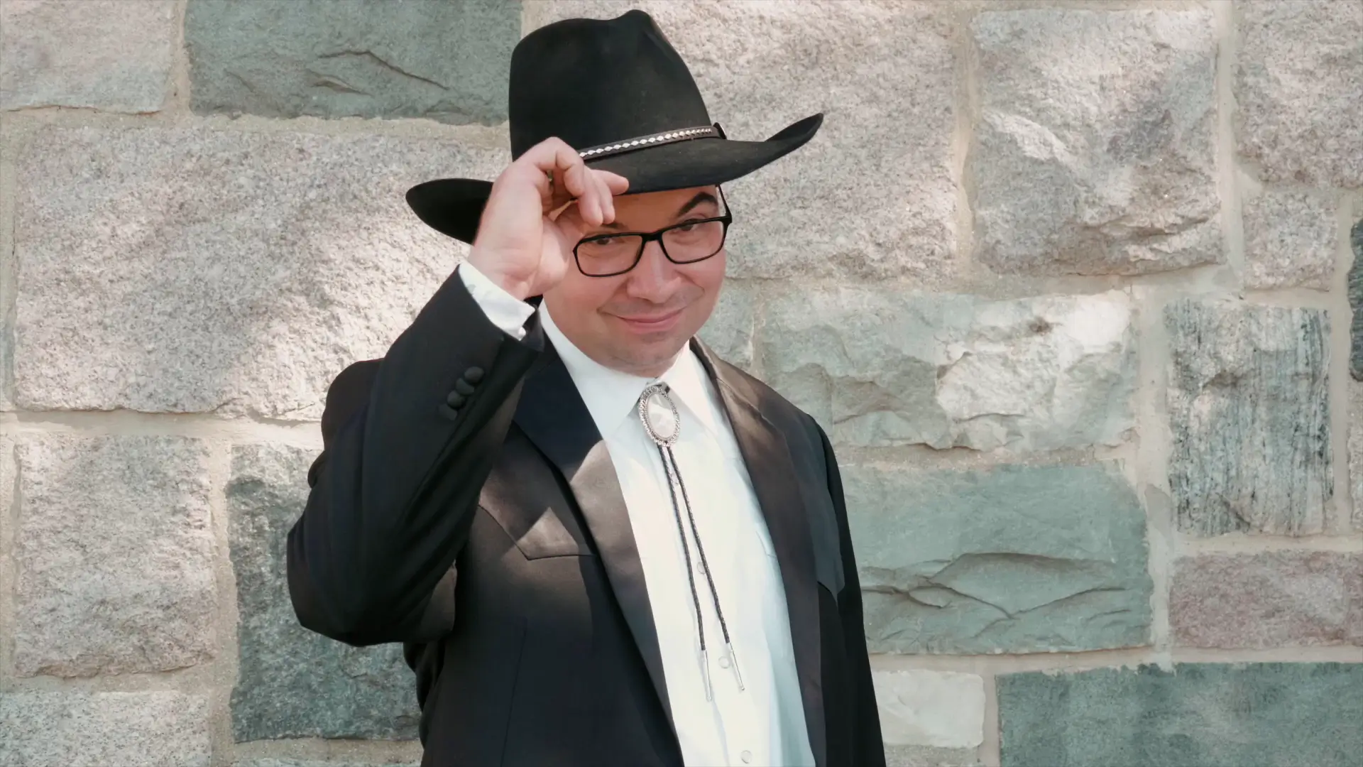 The groom dons his cowboy hat and bolo tie, tipping the hat jovially as he smiles at the videographer's camera against a stone wall backdrop.