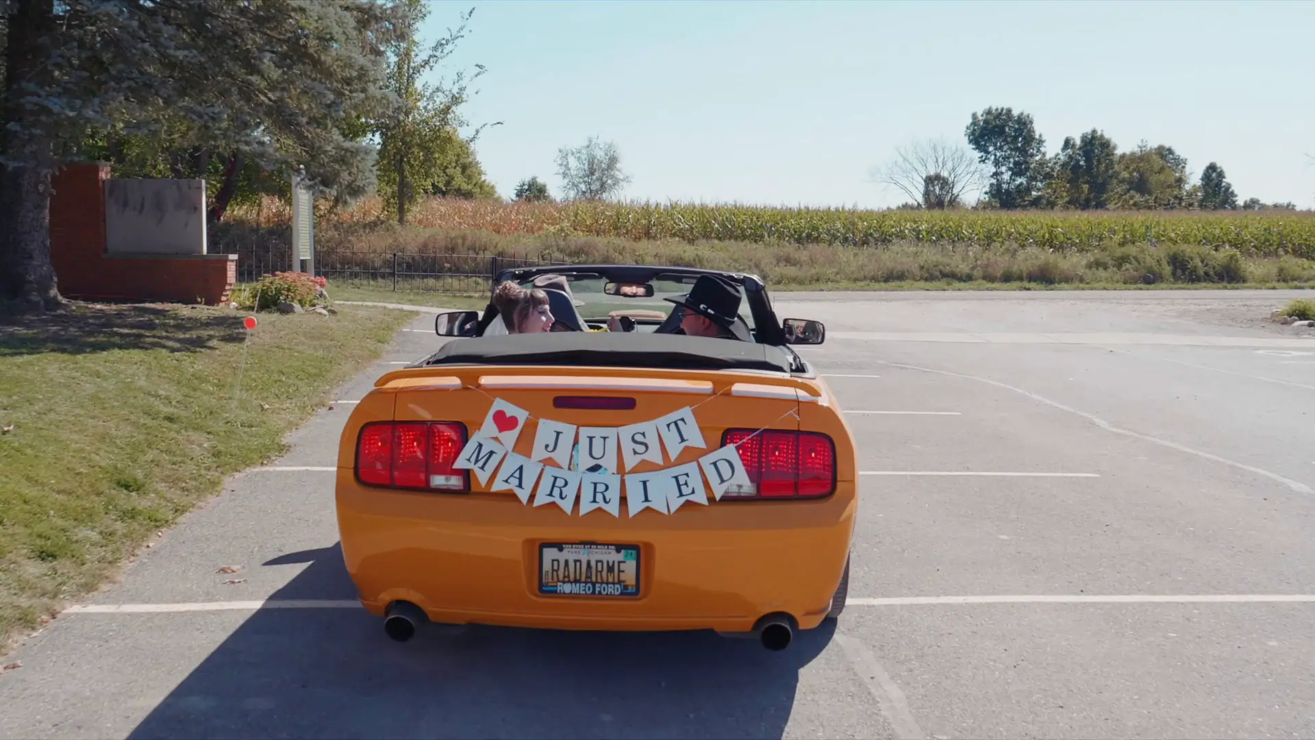 Yellow convertible with "Just Married" sign drives through sunny Michigan countryside, Felicia and Leigh smiling, tall trees and open fields in the background.