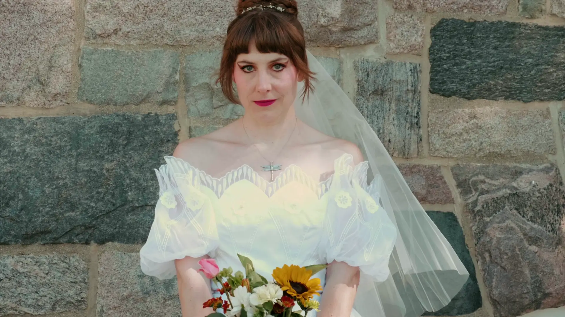 Bride Felicia poses in her wedding dress with a bouquet of wildflowers against a stone wall at St. Augustine Catholic Church, highlighting a serene moment captured by the wedding videographer.