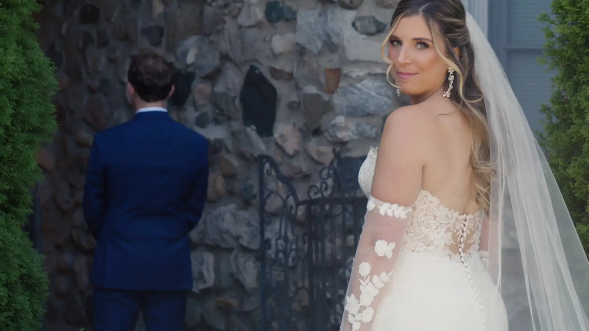 Bride in an elegant lace wedding dress smiles at the camera while standing in front of a stone wall. A groom in a navy suit stands with his back turned, waiting to turn around for their first look.