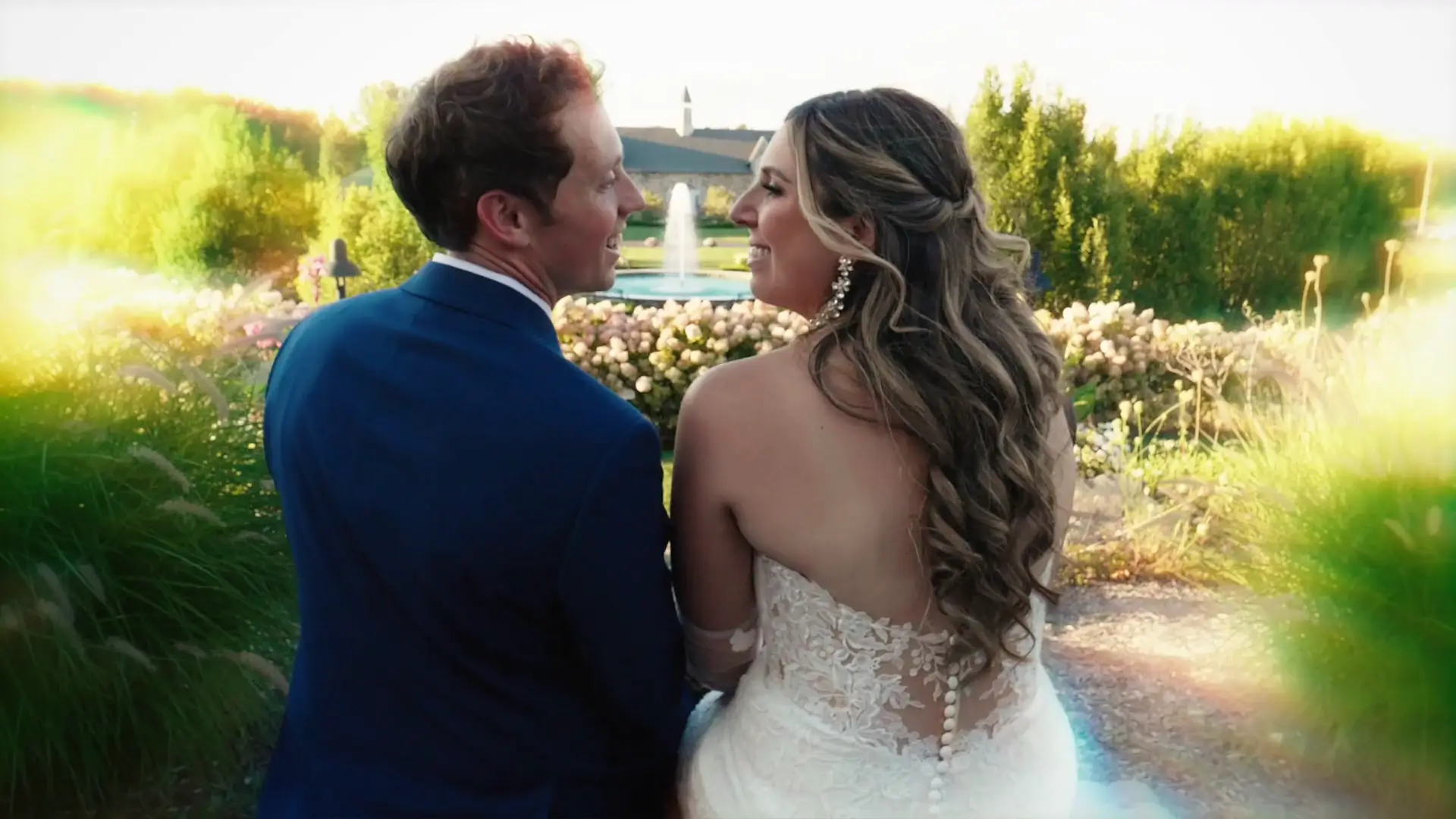 A couple, Jordan and Mads, are seated together, smiling at each other in an outdoor setting at Castle Farms. The bride wears a lace gown, and the groom is in a blue suit. A fountain and lush greenery are visible in the background.