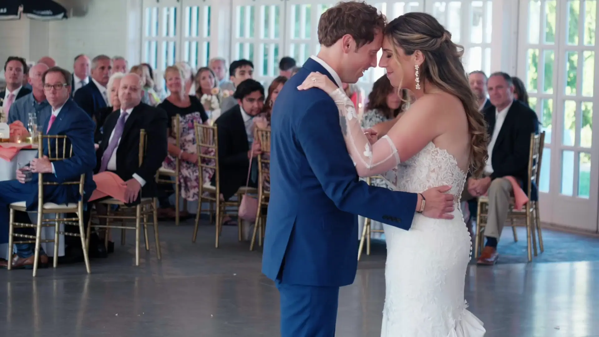 Bride and groom share a tender first dance at their Michigan wedding reception, surrounded by seated guests in the elegant East Garden Room at Castle Farms. The bride wears a lacy white gown, while the groom is in a blue suit, creating a touching moment.
