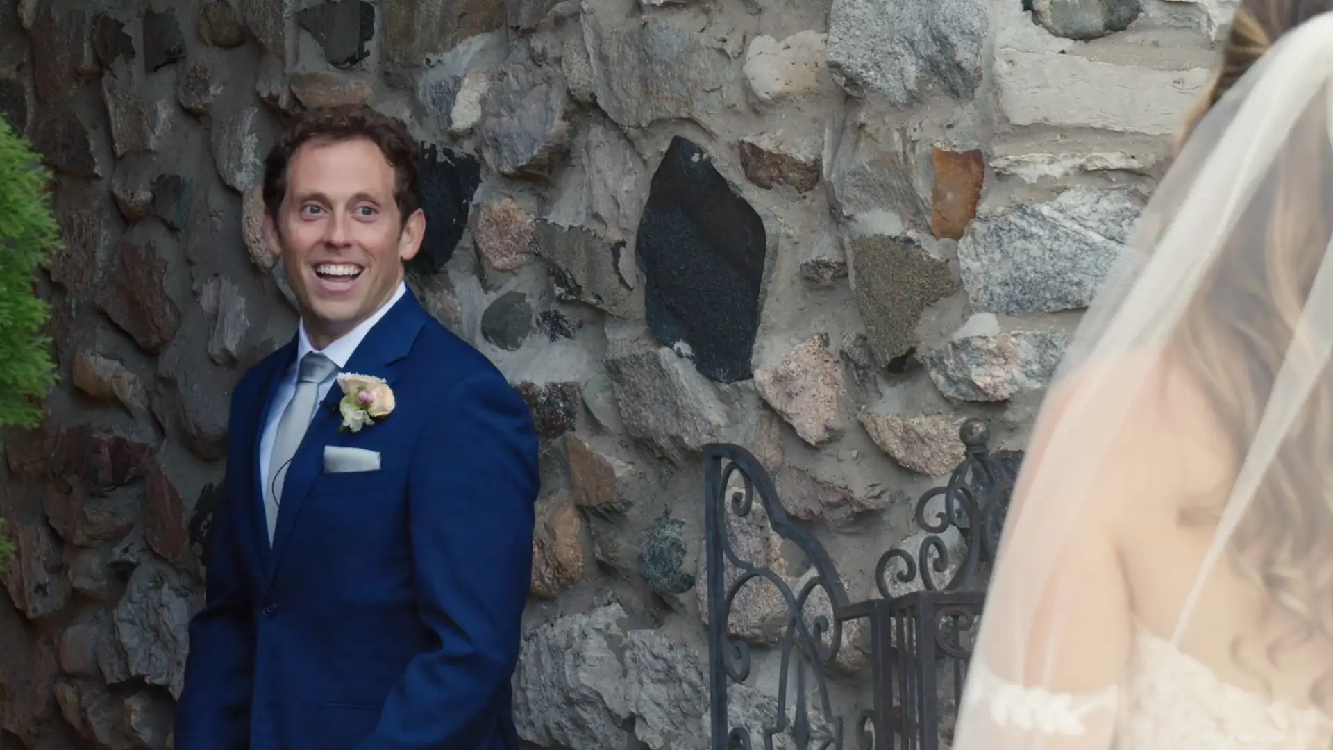 Groom in a blue suit smiling broadly as he beholds his bride during the first look. They stand under a stone archway. The bride's veil and shoulder are visible on the right.