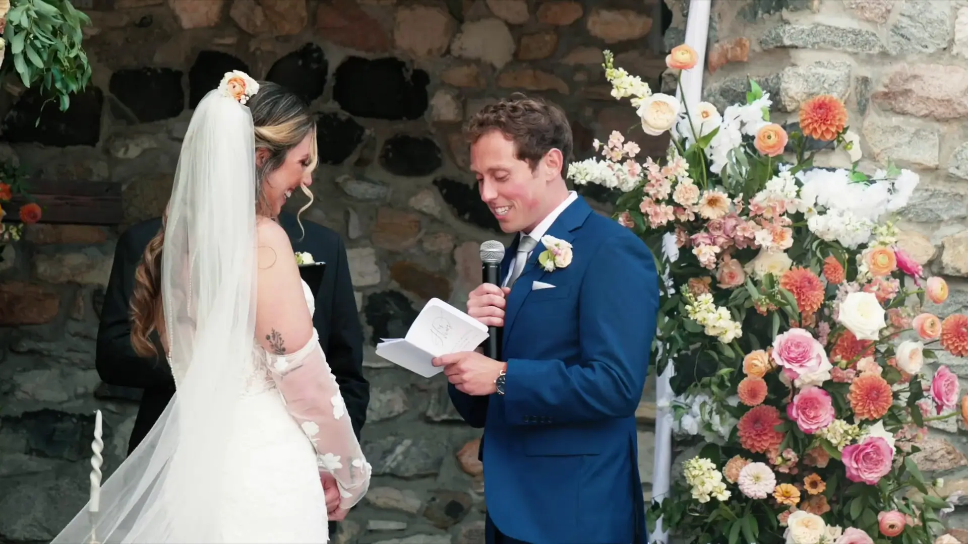 Bride Mads and groom Jordan exchange vows surrounded by vibrant floral arrangements against a stone backdrop at Castle Farms, Michigan. Jordan, in a blue suit, reads his vows as Mads, in a lace gown, smiles lovingly.