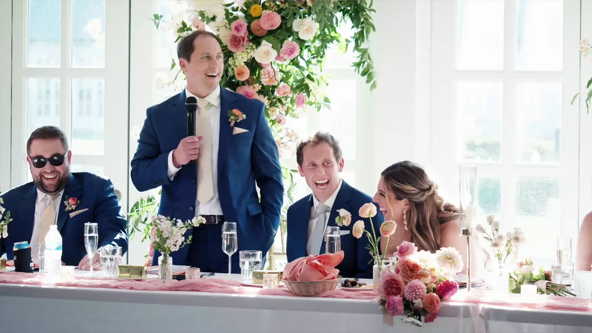 A joyful wedding reception scene at Castle Farms in Michigan, featuring a man giving a speech while the couple and guests smile and laugh. The table is elegantly decorated with flowers, adding to the festive atmosphere.