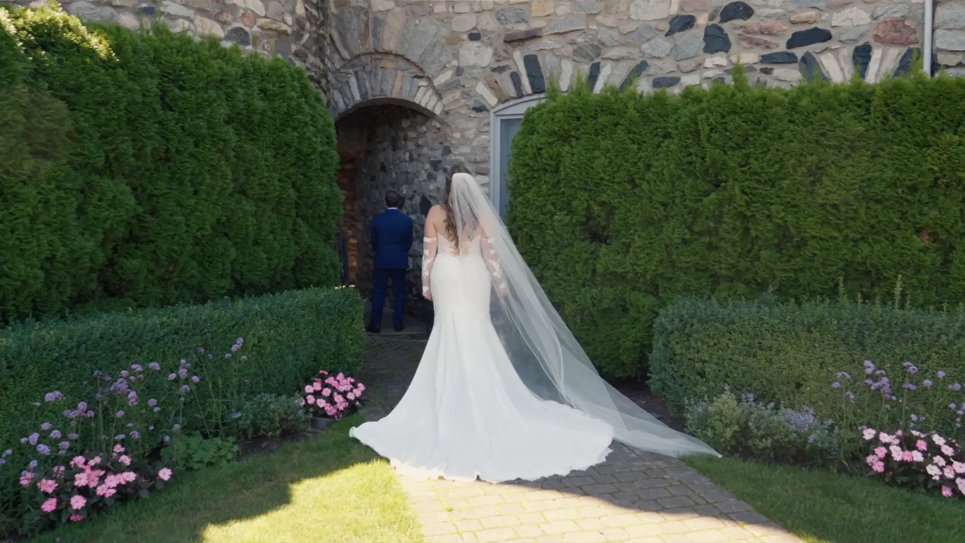 Bride in a flowing white gown with a long veil walks towards a stone archway at Castle Farms, surrounded by lush Michigan greenery and colorful flowers. Under the archway the groom waits in a blue suit for their first look.