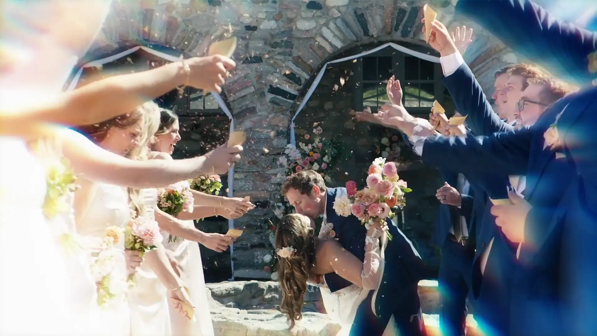 Jordan and Mads kiss amidst cheering friends at their Castle Farms wedding. Bridesmaids and groomsmen in pastel dresses and navy suits throw confetti, forming a lively celebration tunnel. The stone backdrop and floral decor enhance the festive scene.