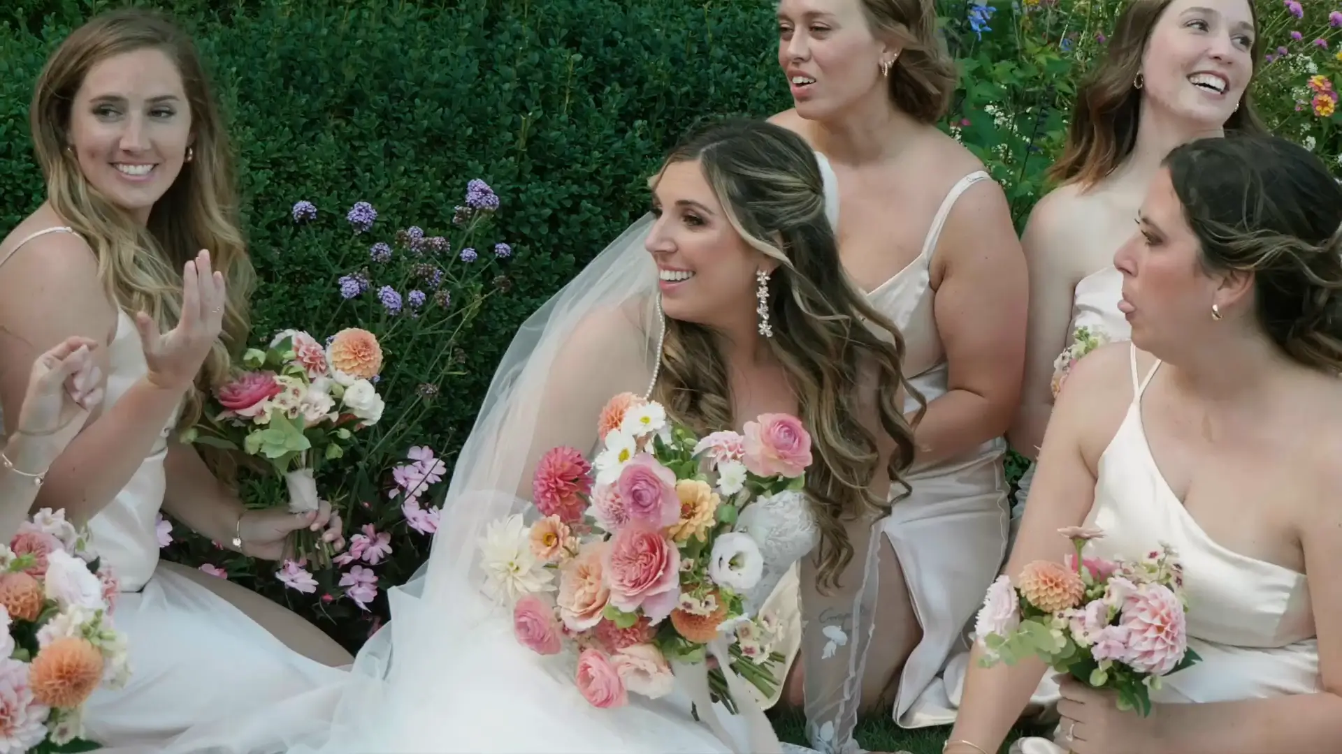 Bridesmaids in elegant dresses and the bride, holding vibrant pink and peach floral bouquets, smile and laugh together amidst lush Michigan greenery at a wedding in Charlevoix, Michigan.