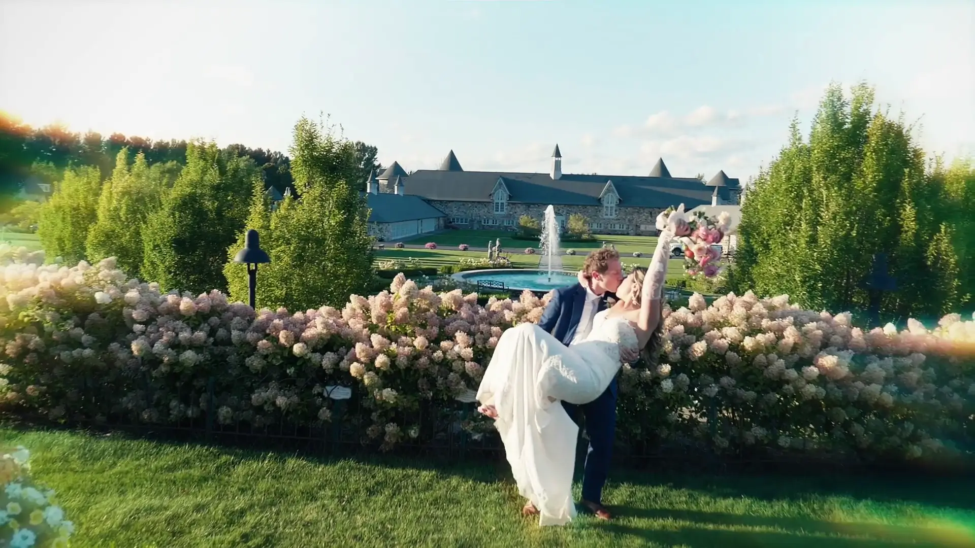A groom in a blue suit lifts the bride in a white gown, holding a bouquet aloft. They are set against a backdrop of lush gardens and stone buildings at Castle Farms in Charlevoix, Michigan.