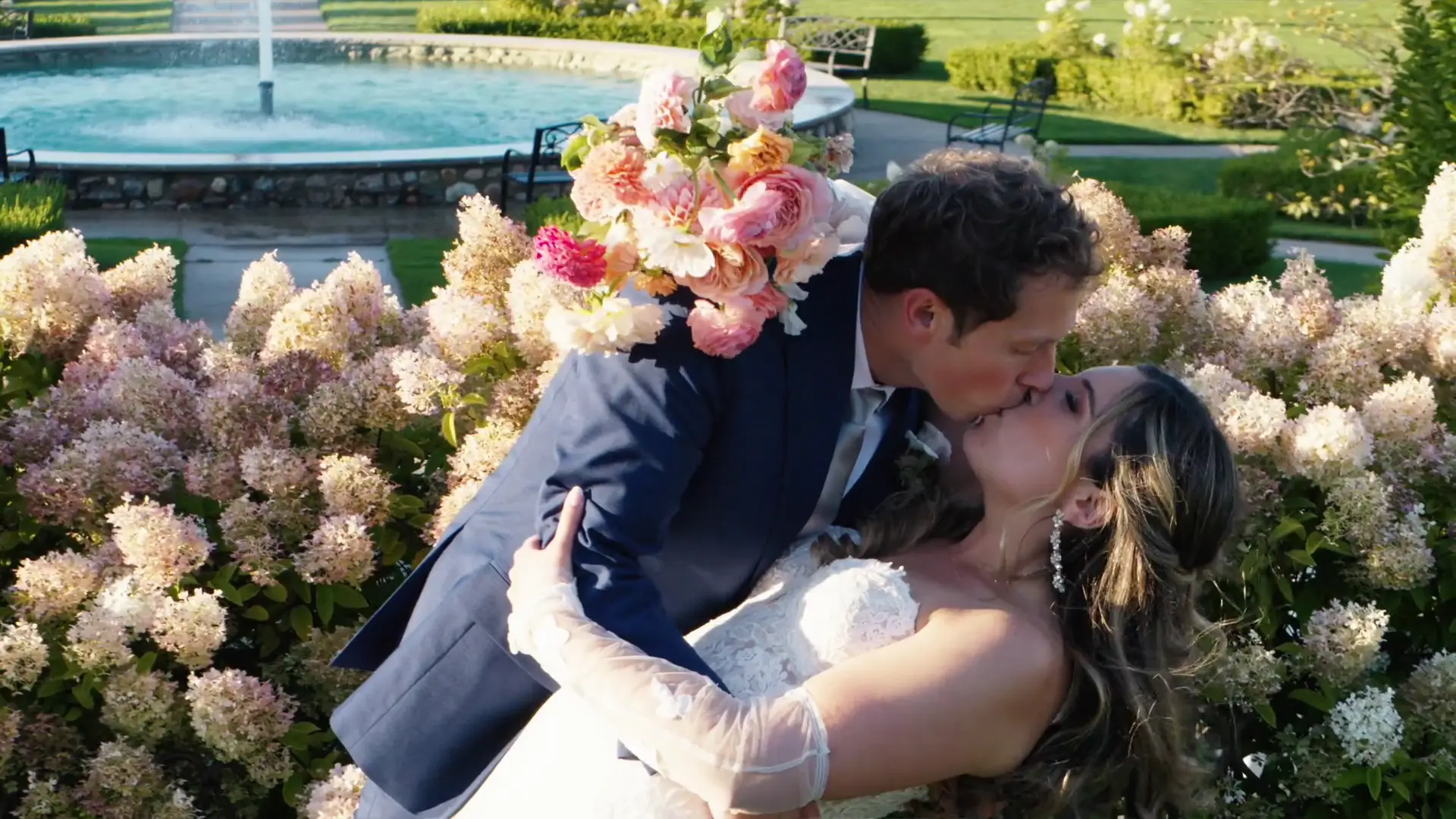 A couple, Jordan and Mads, share a romantic kiss in a garden setting at Castle Farms, Charlevoix, Michigan. The groom dips the bride as she holds a bouquet of pink and peach flowers. A fountain and lush greenery are visible in the background.