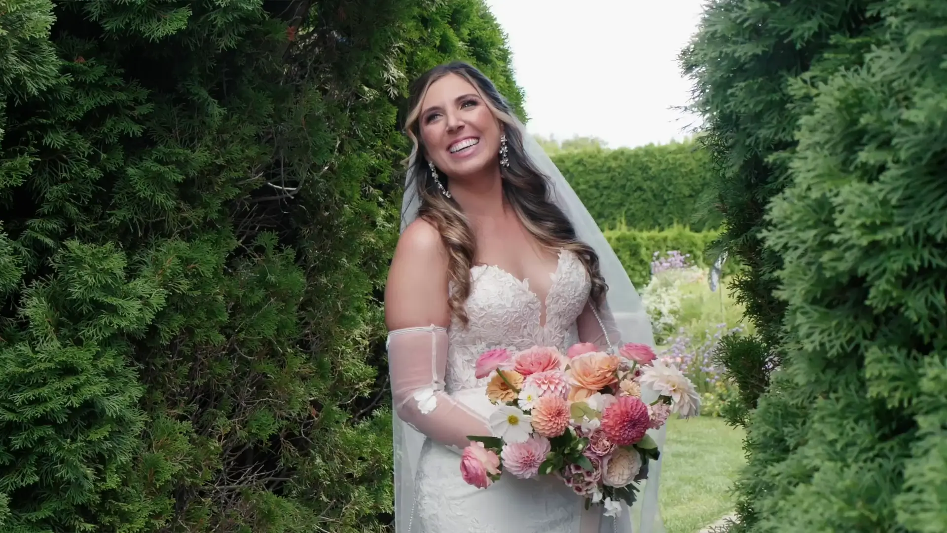 Bride holding a colorful floral bouquet, standing between lush greenery at Castle Farms, Charlevoix. She wears an embroidered lace wedding dress with sheer gloves, smiling warmly during an outdoor photoshoot that reflects the wedding's elegant theme.