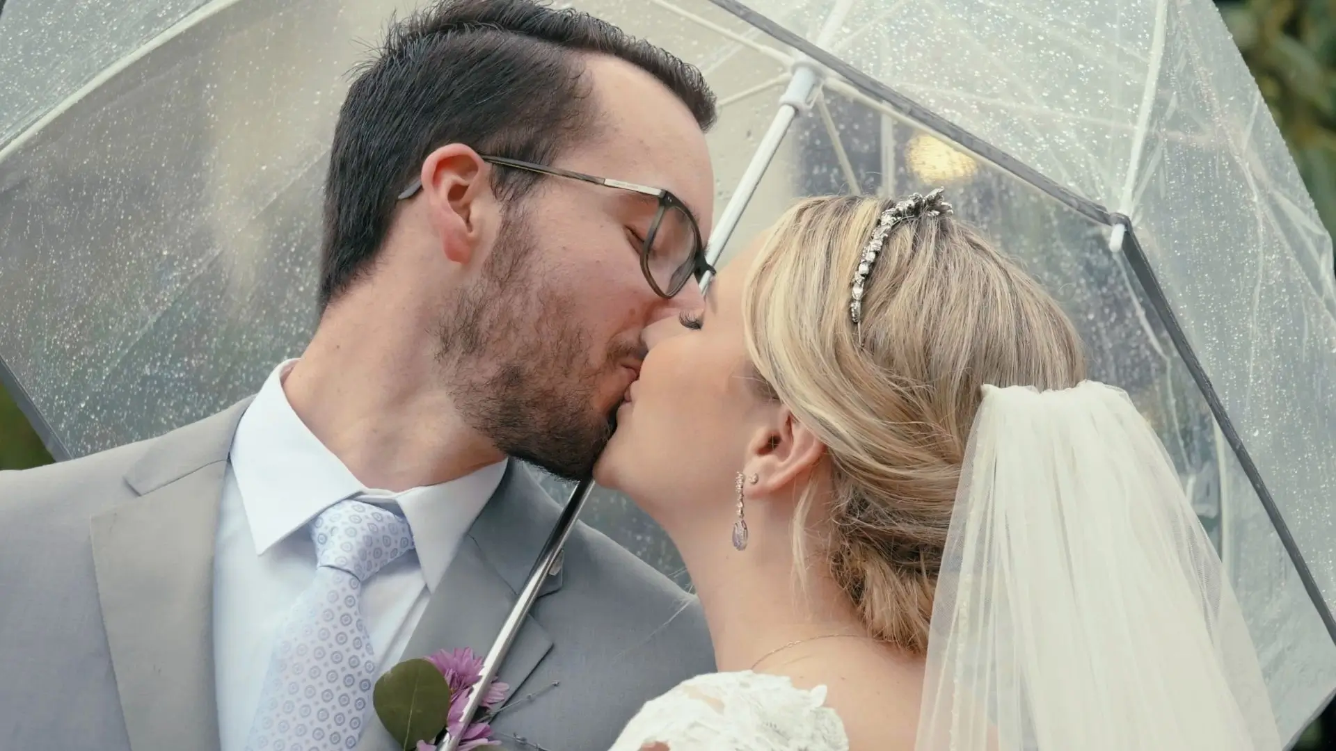 A bride and groom kiss under a clear umbrella on a rainy wedding day at The Union Pavilion in Byron Center, MI. The groom wears a light gray suit.