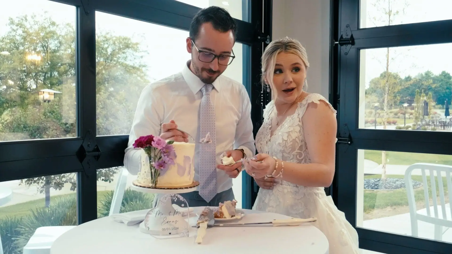 Mckenna and Jacob share a fun moment feeding each other cake in front of large windows at The Union Pavilion at Railside in Byron Center, MI. The table is decorated with purple flowers.